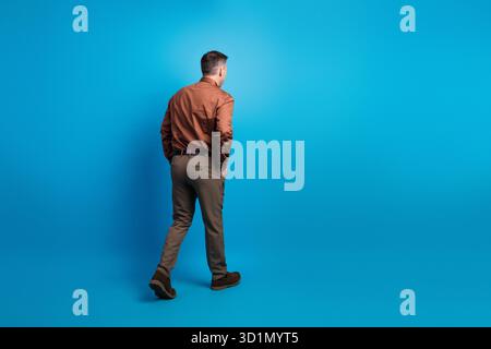 Homme d'affaires professionnel s'éloignant de la caméra dans une chemise brune et un pantalon gris sur un fond de studio bleu vif pour le travail d'entreprise et Banque D'Images
