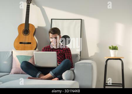 Les jeunes positifs millénaire man sitting on sofa in living room with laptop seul sur week-end à la maison Banque D'Images
