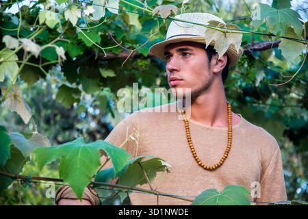 Vue latérale du beau jeune homme en chapeau toching feuilles de vigne dans la lumière du soleil dans le jardin Banque D'Images