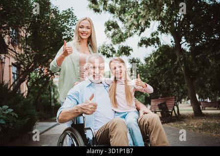 Famille joyeuse dans le parc grand-père en fauteuil roulant avec fille et petite-fille souriant et donnant les pouces Banque D'Images