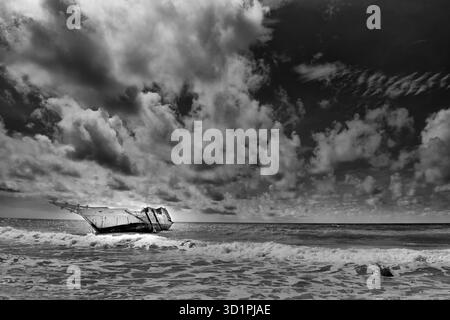 Un naufrage ou un bateau cassé a débarqué dans les vagues d'une mer agitée, photographié en noir et blanc à fort contraste sous un ciel spectaculaire et nuageux Banque D'Images