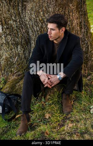 Beau jeune homme se penchant contre l'arbre en automne, portant manteau noir Banque D'Images