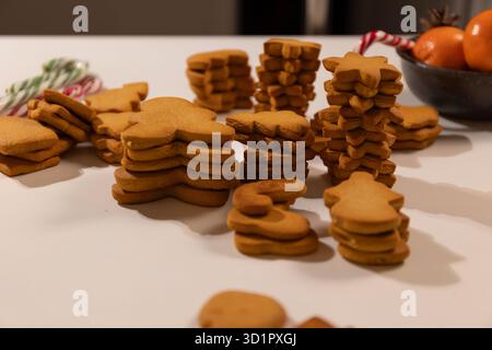 Des biscuits en pain d'épices fraîchement cuits de différentes formes, comme des arbres et des étoiles, sont empilés sur une table blanche aux côtés de cannes de bonbons et de mandarines, prêts pour une célébration de Noël. Banque D'Images