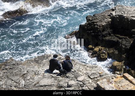 Syrakus, Sizilien - Italie - 04-09-2025 : deux personnes assises sur la rive rocheuse à Syracuse regardant la mer bleue avec des vagues blanches s'écraser contre la côte un Banque D'Images