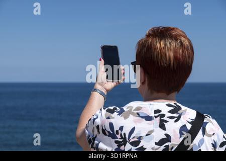 Syrakus, Sizilien - Italie - 04-09-2025 : une femme aux cheveux courts et roux tient un smartphone vers la mer à Syrakus, capturant la vue sous un clair Banque D'Images