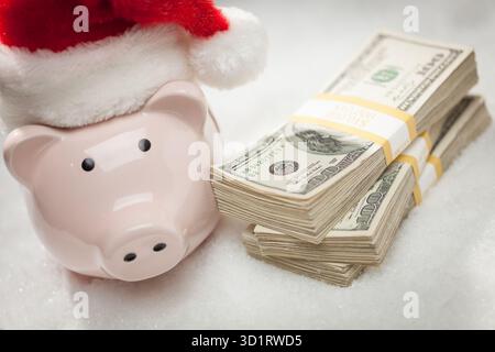 Piggy Bank portant un chapeau de Père Noël près des piles de billets de cent dollars sur des flocons de neige Banque D'Images