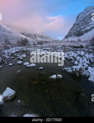 Vue hivernale sur la vallée de Glencoe. La rivière Coe coule sur des rochers poussiéreux de neige, encadrés par d'imposantes montagnes escarpées sous un ciel froid et nuageux au coucher du soleil Banque D'Images