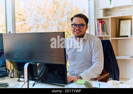 Un directeur d'entreprise, vêtu d'une chemise blanche à la coiffure moderne, se concentre intensément sur son ordinateur dans un cadre de bureau élégant et professionnel. Banque D'Images