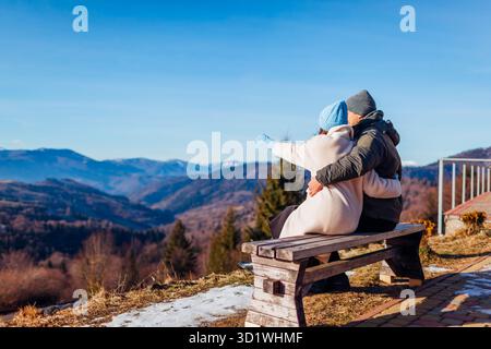 Heureux couple de voyageurs amoureux appréciant le paysage des montagnes des Carpates d'hiver assis sur le banc avec les bras ouverts. Vue enneigée. Vue arrière Banque D'Images