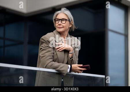 Femme d'affaires âgée dans une tenue élégante se tient sur un balcon tout en tenant son smartphone et en observant le paysage urbain pendant un Banque D'Images