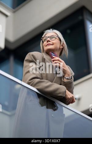 Femme d'affaires âgée regarde avec confiance la ligne d'horizon tout en se tenant debout sur un balcon dans une zone urbaine en fin d'après-midi Banque D'Images