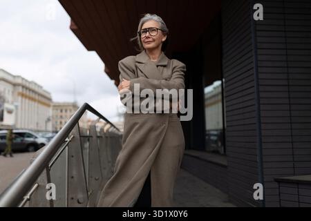 Femme d'affaires âgée se tient en toute confiance devant son bureau, vêtue d'un manteau élégant et de lunettes, par une journée nuageuse dans une ville se Banque D'Images