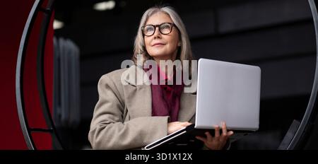 Femme d'affaires âgée se tient devant son bureau tout en tenant un ordinateur portable pendant les heures de lumière du jour Banque D'Images