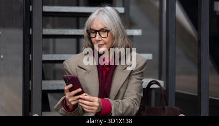 Femme d'affaires d'âge moyen dans un cadre urbain à la mode vérifiant son smartphone tout en étant assise sur les marches par une journée ensoleillée Banque D'Images