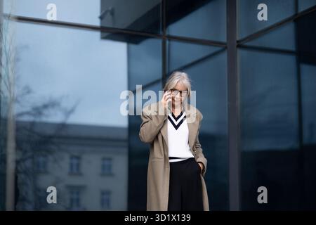 Une femme d'affaires senior en tenue professionnelle parlant sur son téléphone à l'extérieur d'un immeuble de bureaux moderne pendant la journée Banque D'Images