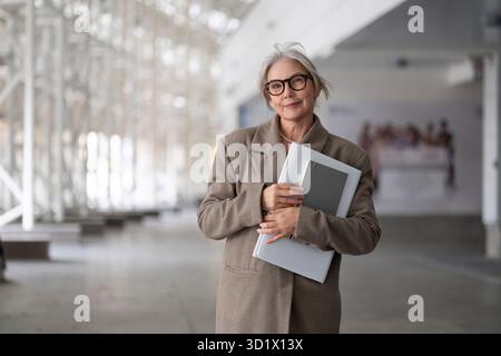 Femme d'affaires senior debout en toute confiance à l'extérieur, tenant un ordinateur portable et présentant son comportement professionnel dans une ville moderne Banque D'Images