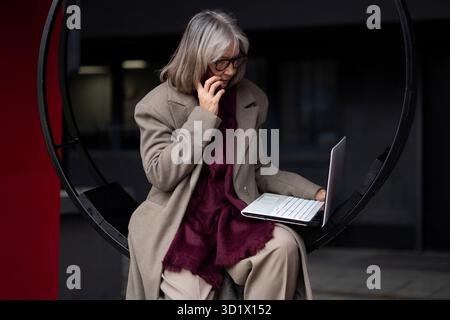 Femme d'affaires âgée dans un manteau élégant parle au téléphone tout en travaillant sur un ordinateur portable dans un cadre extérieur moderne Banque D'Images