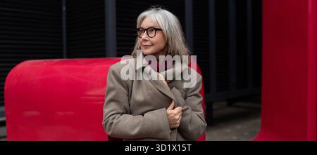 Femme d'affaires âgée se tient en confiance dans un manteau élégant sur un fond rouge vif dans un cadre urbain Banque D'Images