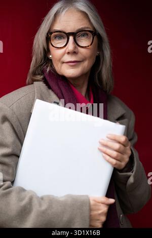 Femme d'affaires caucasienne d'âge moyen tenant un ordinateur portable, posant avec confiance sur un fond rouge vif dans un cadre urbain Banque D'Images
