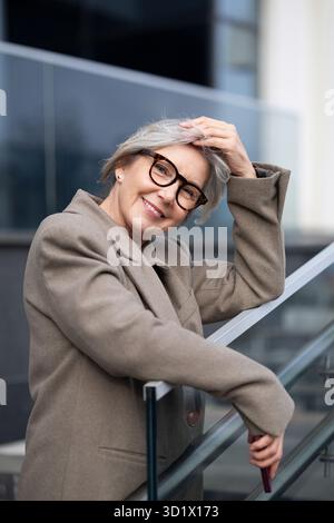 Une femme d'affaires d'âge moyen avec les cheveux gris se tient en toute confiance à l'extérieur, reposant sa main sur une balustrade en verre, souriant chaleureusement dans un env d'entreprise en plein air Banque D'Images