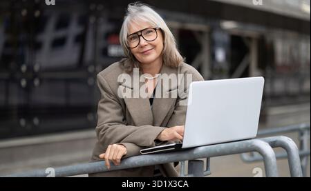Femme plus âgée travaillant sur un ordinateur portable tout en s'appuyant contre une balustrade à l'extérieur dans un cadre urbain par une journée ensoleillée Banque D'Images