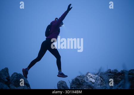 Silhouette d'une femme de fitness coureur de trail sautant par-dessus une falaise et courant à un sommet de montagne rocheuse Banque D'Images