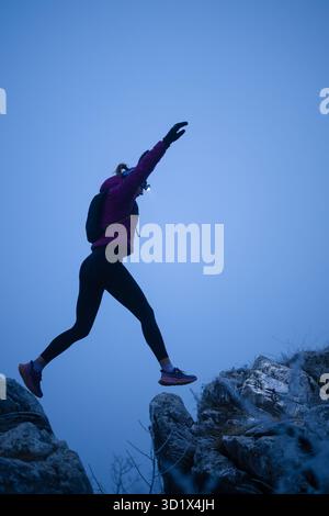 Silhouette d'une femme de fitness coureur de trail sautant par-dessus une falaise et courant à un sommet de montagne rocheuse Banque D'Images