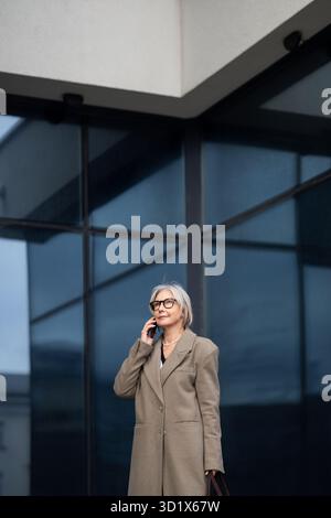 Femme d'affaires âgée dans un manteau élégant parlant sur son téléphone à l'extérieur d'un immeuble de bureaux moderne pendant la journée Banque D'Images