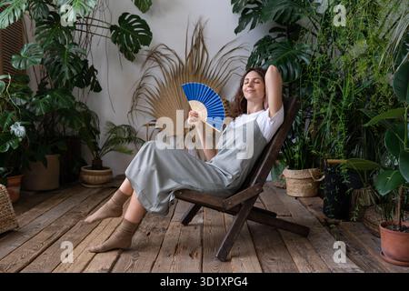 Jeune femme rêveuse et détendue utilisant un ventilateur de papier tout en se relaxant dans le beau jardin rafraîchissant de la maison Banque D'Images