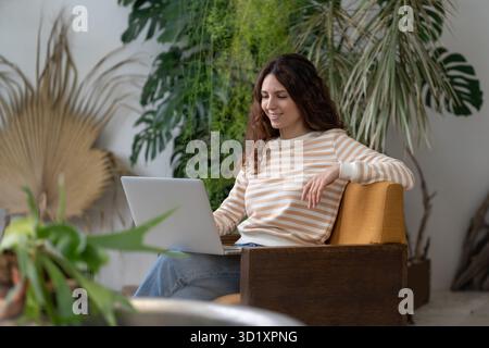 Jeune femme indépendante assise dans un fauteuil en serre, travaillant sur un ordinateur portable entouré d'une maison Banque D'Images