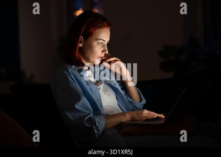 Femme pigiste focalisée regardant l'écran d'ordinateur portable la nuit travaillant de la maison. Banque D'Images