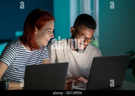 Heureux couple diversifié ont de petites affaires en ligne au travail à la maison sur les ordinateurs portables en soirée Banque D'Images