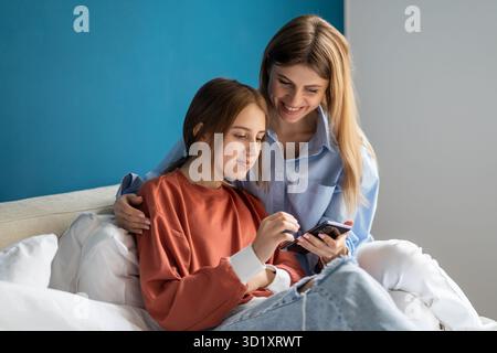 Mère et fille de l'adolescence souriantes et insouciantes utilisant un smartphone ensemble tout en se relaxant dans le lit Banque D'Images