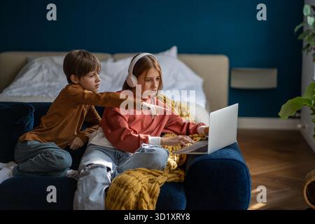Sœur adolescente dans les écouteurs et frère et sœur regardant le film ensemble sur ordinateur portable assis sur le canapé à la maison. Garçon pointant quelque chose sur l'écran. Le Banque D'Images