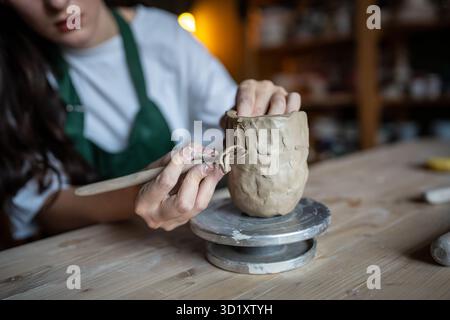 Les mains féminines utilisent habilement l'outil de boucle de poterie pour gratter l'argile et sculpter le motif décoratif sur la tasse Banque D'Images