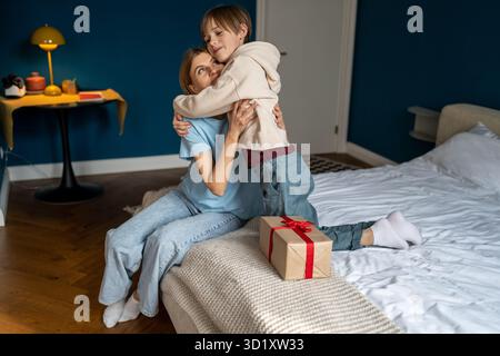Maman donnant cadeau surprise sur l'anniversaire de son fils assis dans le lit à la maison, garçon embrassant la femme. Banque D'Images