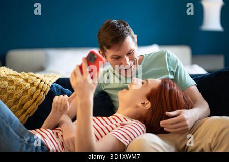 Homme allemand et femme avec smartphone riant à des vidéos drôles Internet couché sur le canapé. Banque D'Images