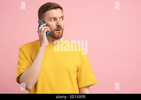 L'homme d'étonnement regarde loin dans la conversation surprise sur smartphone isolé sur l'espace de copie de fond rose Banque D'Images