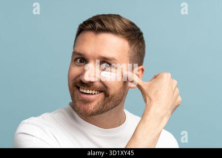 L'homme heureux souriant applique une crème hydratante sur le visage avec un effet anti-âge sur fond de studio Banque D'Images