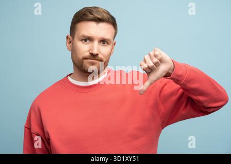 Homme irrité mécontent montrant pouce vers le bas geste d'aversion isolé sur fond bleu studio. Banque D'Images