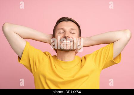 Homme insouciant souriant avec les yeux fermés mains détendues au-dessus de la tête isolé sur fond rose espace copie Banque D'Images