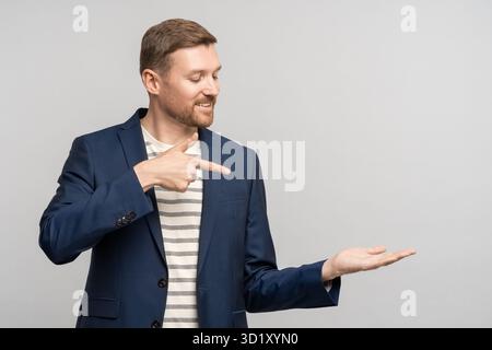 Homme d'affaires barbu heureux souriant pointant l'index sur la main sur l'espace de copie sur fond gris. Banque D'Images