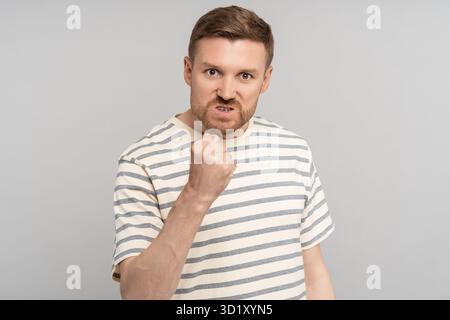 Homme en colère montrant le poing avec grimace mécontente et yeux fous regardant la caméra isolée sur le mur gris Banque D'Images