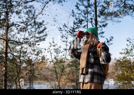 Observateur d'oiseaux touristique de femme concentrée dans la forêt d'automne regardant à travers des jumelles sur les oiseaux. Tourisme. Banque D'Images