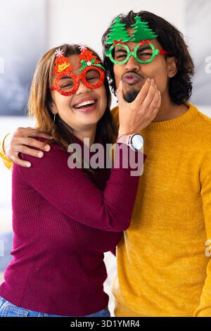Couple diversifié embrassant et riant tout en portant des lunettes de Noël et smartwatch dans le salon Banque D'Images