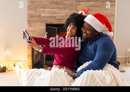 Couple divers portant des chapeaux couverture blottissante dans le salon par cheminée posant smartphone selfie Banque D'Images