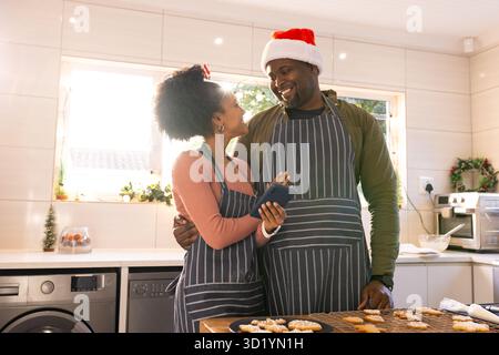Couple afro-américain portant des tabliers serrant dans la cuisine tout en vérifiant smartphone par plateau à biscuits Banque D'Images