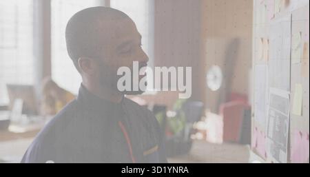Homme portant une chemise sombre examinant les plans au mur de briques dans le bureau, avec des notes adhésives et des graphiques Banque D'Images
