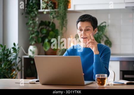 Gestes engagés de femme d'affaires mature expliquant les tâches sur la réunion vidéo à la maison avec du thé et des notes Banque D'Images