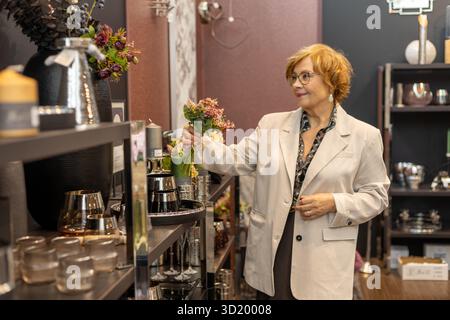Une femme aux cheveux bouclés et aux lunettes examine des objets décoratifs dans une boutique de décoration chic. Elle tient un petit bouquet tout en sélectionnant les pièces parfaites Banque D'Images